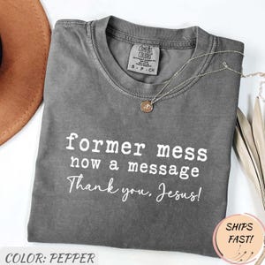 Könnte beinhalten: Ein pfefferfarbenes Comfort Colors T-Shirt mit dem weißen Text "former mess now a message Thank you, Jesus!". Das Shirt hat einen Rundhalsausschnitt und kurze Ärmel. Eine silberne Halskette mit Anhänger liegt auf dem Shirt.
