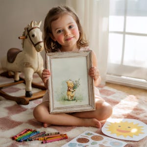 May include: A young girl sits on a pink and white rug, holding a framed picture of Winnie the Pooh. The picture has a distressed white frame. A wooden rocking horse and crayons are also visible.