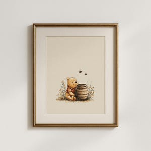May include: Framed watercolor print featuring a seated yellow bear in a red shirt, next to a brown honey pot. Two bees fly above the pot. The artwork is set against a cream background, framed in a gold-colored frame.