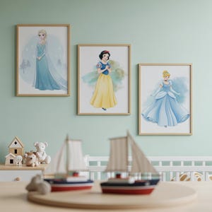 Puede incluir: Tres impresiones art&iacute;sticas enmarcadas con princesas de Disney: Elsa con un vestido azul, Blancanieves con un vestido amarillo y Cenicienta con un vestido de gala azul. Las impresiones est&aacute;n en una pared verde claro, con veleros de juguete y una casa de juguete de madera en primer plano.