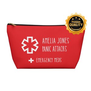Puede incluir: Una bolsa roja con cremallera con texto blanco y un símbolo médico. El texto dice "AMELIA JONES PANIC ATTACKS EMERGENCY MEDS". Un sello dorado de "Premium Quality" está en la esquina superior derecha.