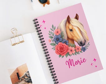 May include: A spiral-bound notebook with a pink cover featuring a watercolor illustration of a horse's head surrounded by flowers. The name "Morie" is written in cursive. The notebook is open on a white surface.