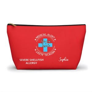 Può includere: Un astuccio rosso con cerniera e cerniera e finiture nere. L'astuccio presenta un design di allerta medica con una croce blu e le parole "Epi pen Inside". Include anche il testo "Severe Shellfish Allergy" e il nome "Sophia".