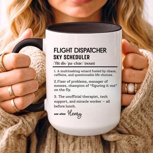May include: A white ceramic mug with a black handle and rim, featuring the text "FLIGHT DISPATCHER SKY SCHEDULER" and a definition. The definition describes the role with humorous statements. The mug is held by a person wearing a beige sweater.