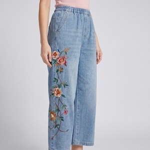 May include: Light blue denim trousers featuring an elasticated waistband and a wide-leg silhouette. The front of the left leg is decorated with an intricate, colourful floral embroidery pattern in shades of red, orange, pink, and green.