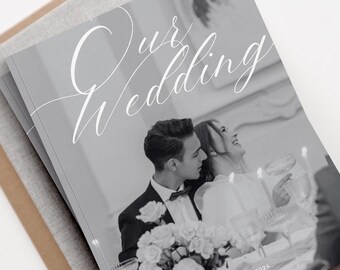 May include: A stack of wedding photo albums with the words "Our Wedding" in elegant script. The cover is a light gray color, and the image shows a couple at a wedding reception. The album is resting on a neutral-toned surface.