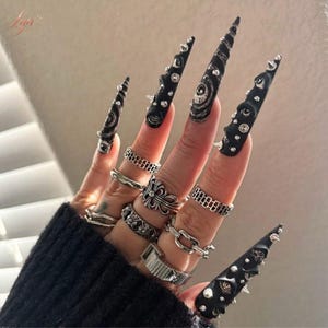 Könnte beinhalten: Eine Hand, die lange, schwarze Stilettnägel mit silbernen Nieten und wirbelnden weißen Designs zeigt. Die Finger sind mit verschiedenen silbernen Ringen geschmückt, darunter Ketten-, Blumen- und Bandstile. Die Nägel haben eine gotische Ästhetik.