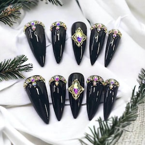 Puede incluir: Un conjunto de diez uñas postizas negras brillantes en forma de estilete. Cada uña está adornada con pedrería iridiscente, algunas con una joya central en forma de diamante en un engaste dorado. Las uñas están dispuestas sobre una superficie blanca.