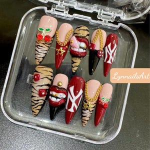 Pode incluir: Um conjunto de dez unhas artificiais numa caixa de plástico transparente. As unhas apresentam vários designs, incluindo riscas de tigre, detalhes de cereja, correntes douradas e um design vermelho e branco. As unhas são de várias formas e cores.