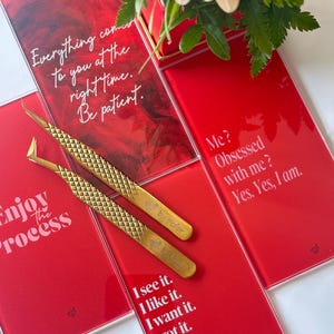 Op de afbeelding: Een close-up van gouden pincetten en rode kaarten met witte tekst. De kaarten tonen zinnen als "Geniet van het proces" en "Ik zie het. Ik vind het leuk. Ik wil het. Ik heb het." Een boeket bloemen staat op de achtergrond.