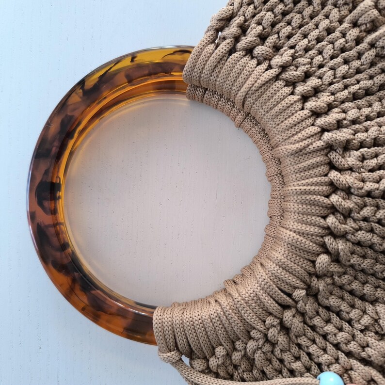 Puede incluir: Primer plano de un bolso de mano de macram&eacute; beige con un asa de carey. La textura tejida del bolso es visible, y una peque&ntilde;a cuenta azul cuelga de la parte inferior. El asa tiene una forma circular grande con tonos marrones y &aacute;mbar.