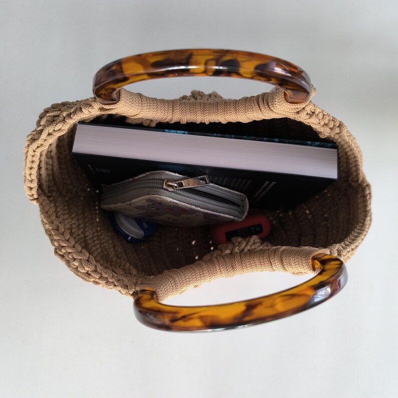 Puede incluir: Un bolso de mano tejido en color beige con asas de carey. El bolso est&aacute; abierto, revelando un libro, una peque&ntilde;a cartera y otros objetos. Las asas son curvas y est&aacute;n hechas de un material brillante y estampado.