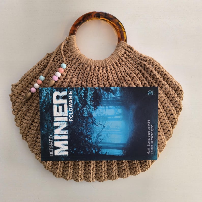 Puede incluir: Un bolso de mano tejido en crochet color beige con un asa de carey, que contiene un libro titulado "MINIER POLOWANIE". El bolso tiene un cierre de cord&oacute;n con cuentas de colores pastel. La portada del libro presenta una escena de bosque azul.
