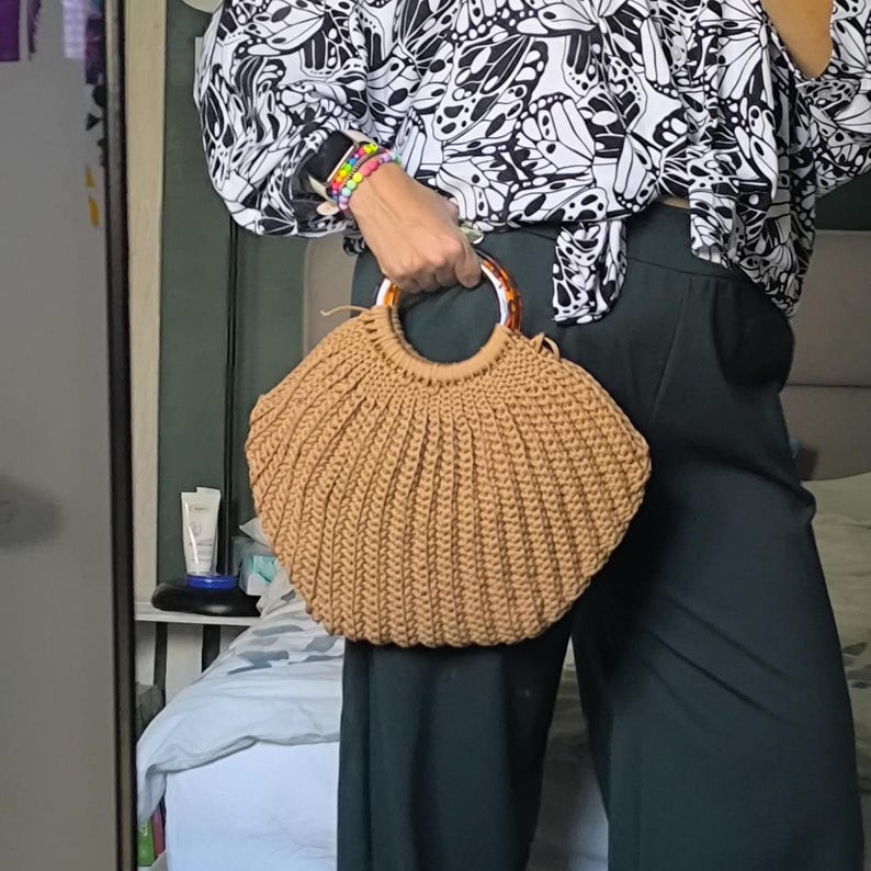 Puede incluir: Un bolso de mano tejido a mano de color marr&oacute;n claro, con un asa circular con estampado de carey. El bolso presenta un dise&ntilde;o plisado y una forma redondeada. El bolso lo sostiene una persona que lleva una blusa negra y blanca con estampado de mariposas y pantalones verde oscuro.