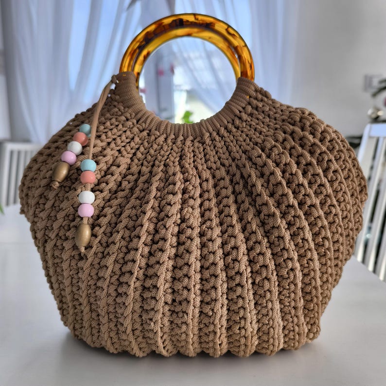 Puede incluir: Un bolso de mano tejido a mano en color beige, con forma redondeada y asa de carey. El bolso presenta un dise&ntilde;o acanalado y texturizado y una borla decorativa con cuentas de colores pastel. El bolso est&aacute; sobre una superficie blanca.