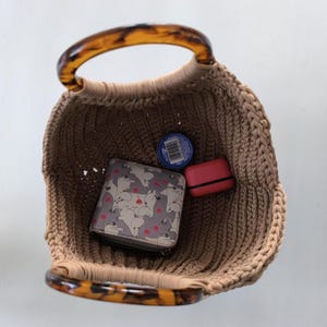 Puede incluir: Un bolso tote tejido de color beige con asas de carey. Dentro de la bolsa hay una cartera con un estampado de gatos, un estuche rojo peque&ntilde;o y un recipiente azul.