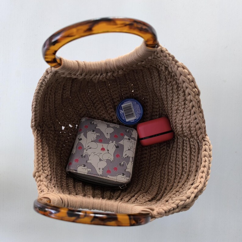 Puede incluir: Un bolso tote tejido de color beige con asas de carey. Dentro de la bolsa hay una cartera con un estampado de gatos, un estuche rojo peque&ntilde;o y un recipiente azul.