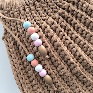 Puede incluir: Primer plano de un bolso de ganchillo color beige con cierre de cord&oacute;n. El cord&oacute;n presenta una serie de cuentas de colores pastel en azul, rosa y blanco, intercaladas con cuentas de madera. La textura del bolso es visible.