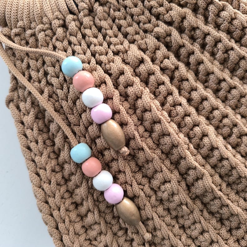 Puede incluir: Primer plano de un bolso de ganchillo color beige con cierre de cord&oacute;n. El cord&oacute;n presenta una serie de cuentas de colores pastel en azul, rosa y blanco, intercaladas con cuentas de madera. La textura del bolso es visible.