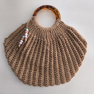 Puede incluir: Bolso de mano de macram&eacute; marr&oacute;n con dise&ntilde;o de concha. Cuenta con un asa con estampado de carey y una borla con cuentas pastel. El bolso est&aacute; hecho de un material grueso y tejido.