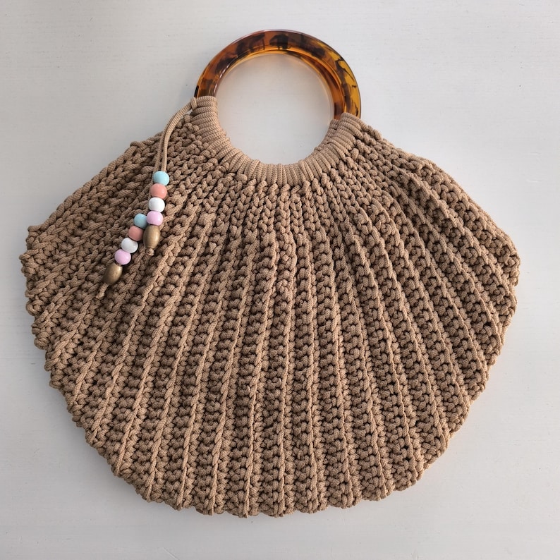 Puede incluir: Bolso de mano de macram&eacute; marr&oacute;n con dise&ntilde;o de concha. Cuenta con un asa con estampado de carey y una borla con cuentas pastel. El bolso est&aacute; hecho de un material grueso y tejido.