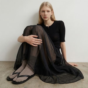 May include: A woman wearing a black short-sleeved ribbed top, a sheer black skirt, and black flip-flops. The skirt is long and flowing, and the flip-flops have black straps. The woman is sitting on a light-colored floor with a white wall in the background.