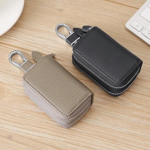 May include: Two leather key cases, one taupe and one black, with silver-tone metal hardware. Each case has a zipper closure and a metal clasp for attaching to a bag or belt loop. The cases are rectangular and appear to be made of textured leather.