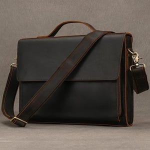 May include: A black leather messenger bag with a top handle and adjustable shoulder strap. The bag features a flap closure and metal hardware. The leather has a distressed appearance, with brown accents on the edges and strap.