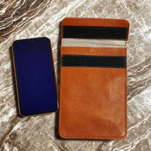 Puede incluir: Una funda de teléfono de cuero naranja con cierres de velcro negros, junto a un smartphone con pantalla azul y marco rojo. La funda está abierta, revelando un interior de color claro. El fondo es una superficie similar al mármol.