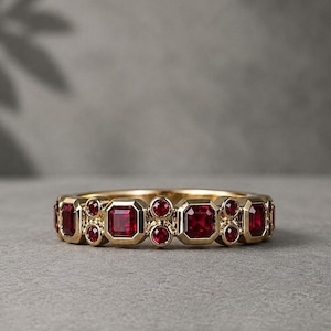 Könnte beinhalten: Ein goldener Ring mit einer Reihe von quadratischen und runden roten Edelsteinen. Die Edelsteine sind in einem sich wiederholenden Muster um das Band angeordnet. Der Ring wird auf einer grauen Oberfläche präsentiert.