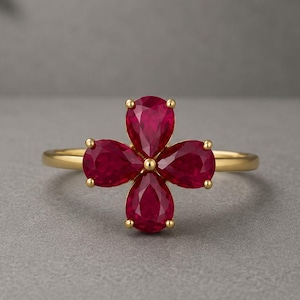 Könnte beinhalten: Ein Goldring mit vier birnenförmigen, tiefroten Rubinen, die in einer blütenartigen Anordnung gruppiert sind. Die Steine sind in einer zentralen Krappenfassung befestigt und bilden ein symmetrisches Blumenmuster auf einem dünnen, polierten Goldring.