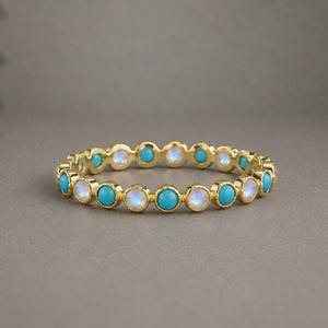 Könnte beinhalten: Ein goldener Ring mit einem abwechselnden Muster aus runden, in Zargenfassung gefassten Türkissteinen und irisierenden Mondsteinen. Die Steine sind in einem durchgehenden Kreis um das Metallband angeordnet, was ein einheitliches Design ergibt.