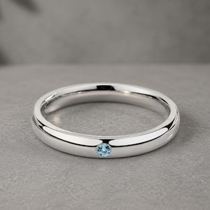 Könnte beinhalten: Ein polierter Silberring mit einem einzelnen, kleinen, runden, hellblauen Edelstein. Der Ring hat ein glattes, abgerundetes Band und ein schlichtes, elegantes Design. Der Edelstein ist in der Mitte des Rings gefasst.