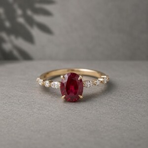 Könnte beinhalten: Ein goldener Ring mit einem großen, oval geschliffenen Rubin als Mittelstein. Das Band ist mit kleineren Diamanten in runden und navetteförmigen Schliffformen verziert. Der Ring ist vor einem neutralen grauen Hintergrund platziert.