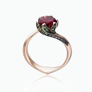 Könnte beinhalten: Roségoldener Ring mit einem großen, runden, tiefroten Edelstein. Die Fassung umfasst grüne und schwarze Akzentsteine mit einem fließenden, organischen Design. Der Ring hat einen elegant geschwungenen Ring.