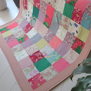 Może przedstawiać: Narzuta typu patchwork składająca się z siatki kwadratowych kawałków materiału w różnych kolorach, w tym różowym, zielonym, czerwonym i białym. Narzuta łączy jednolite kolory z wzorami kwiatowymi lub paisley i jest wykończona jasnoróżową lamówką.