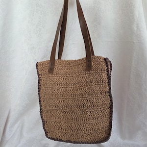May include: A rectangular tote bag crafted from tan woven straw material. The bag features two long brown faux leather shoulder straps attached with dark stitching. The edges of the bag are finished with a dark brown crocheted border trim.