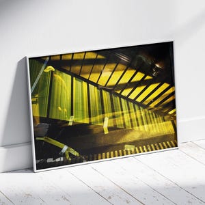 May include: Abstract art print with a yellow and black geometric design. The artwork is framed in a white border and leans against a white wall and floor. The image has a modern aesthetic with strong shadows and light.