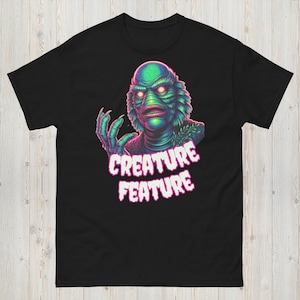 Pode incluir: Camiseta preta com uma ilustração colorida de uma criatura com olhos brilhantes e o texto "CREATURE FEATURE" em rosa e branco. O design tem um tema de filme de terror vintage.