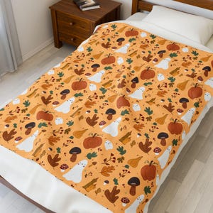 Puede incluir: Una manta naranja con temática de Halloween con un patrón repetido de calabazas, fantasmas, setas y hojas de otoño. La manta está extendida sobre una cama, mostrando el diseño de inspiración otoñal. La manta está hecha de un material suave.