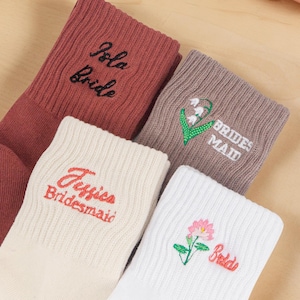 Può includere: Un assortimento di calzini colorati con ricami e motivi floreali. I calzini sono in tonalità di rosso, beige, marrone e bianco. I testi includono "Jola Bride", "Jessica Bridesmaid", "Brides Maid" e "Bride". Una bobina di filo rosa sullo sfondo.