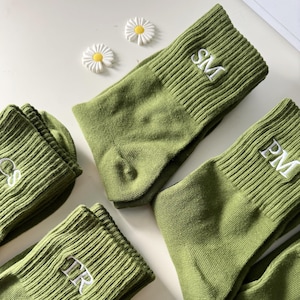 以下が含まれることがあります： 白の刺繍イニシャルが入ったオリーブグリーンの靴下。 靴下はリブ編みで折り返されており、数足のペアが見えます。 白と黄色の小さなデイジーの装飾も2つあります。
