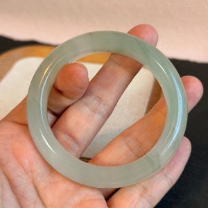 K&ouml;nnte beinhalten: Ein hellgr&uuml;nes Jade-Armreif-Armband. Das glatte, kreisf&ouml;rmige Armband wird in einer Hand gehalten und zeigt seine polierte Oberfl&auml;che und seine durchscheinende Qualit&auml;t. Der Jade weist subtile Farbvariationen auf, die dem Design Tiefe verleihen. Ein klassisches Schmuckst&uuml;ck.
