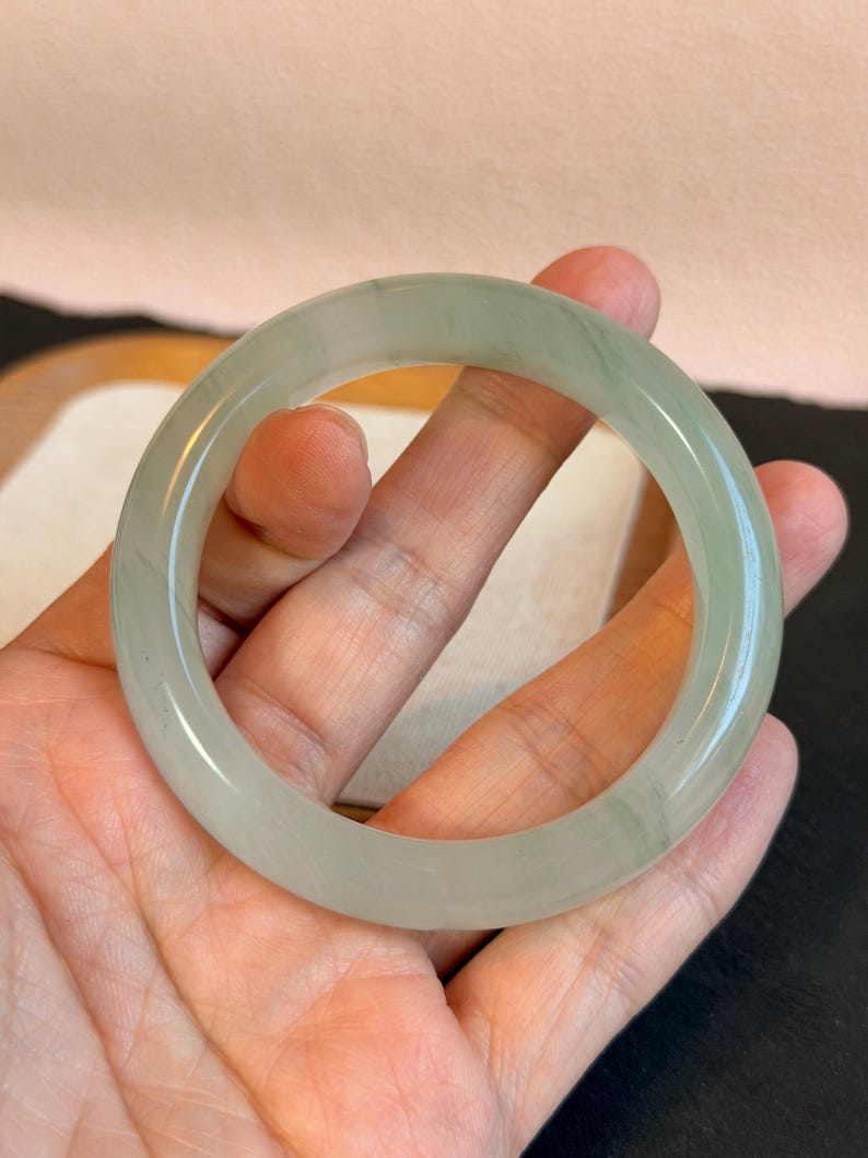 K&ouml;nnte beinhalten: Ein hellgr&uuml;nes Jade-Armreif-Armband. Das glatte, kreisf&ouml;rmige Armband wird in einer Hand gehalten und zeigt seine polierte Oberfl&auml;che und seine durchscheinende Qualit&auml;t. Der Jade weist subtile Farbvariationen auf, die dem Design Tiefe verleihen. Ein klassisches Schmuckst&uuml;ck.