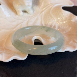 K&ouml;nnte beinhalten: Ein durchscheinendes Jade-Armreif-Armband in blassgr&uuml;ner Farbe. Die polierte Oberfl&auml;che reflektiert das Licht und zeigt nat&uuml;rliche Maserungen. Das Armband wird auf einer wei&szlig;en, blattf&ouml;rmigen Schale mit einer kleinen wei&szlig;en Hasenfigur im Hintergrund pr&auml;sentiert.