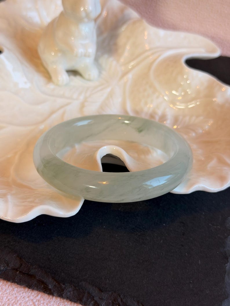 K&ouml;nnte beinhalten: Ein durchscheinendes Jade-Armreif-Armband in blassgr&uuml;ner Farbe. Die polierte Oberfl&auml;che reflektiert das Licht und zeigt nat&uuml;rliche Maserungen. Das Armband wird auf einer wei&szlig;en, blattf&ouml;rmigen Schale mit einer kleinen wei&szlig;en Hasenfigur im Hintergrund pr&auml;sentiert.