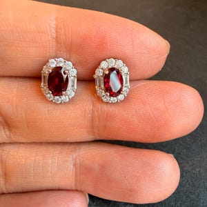 May include: A pair of oval-shaped stud earrings. Each earring features a deep red gemstone surrounded by a halo of small, clear, round and baguette-cut stones. The earrings are set in a silver-colored metal.