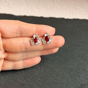 May include: A pair of stud earrings featuring oval-shaped deep red gemstones set in textured silver-toned metal settings. The metal base has an organic, petal-like shape that surrounds the central red stone, creating a distinct contrast between the smooth, polished surface of the gemstone and the rough, detailed finish of the metal frame.