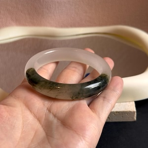 Könnte beinhalten: Ein durchscheinendes Jade-Armreif-Armband. Das Armband ist hauptsächlich ein blasses, milchiges Weiß mit dunkelgrünen Einschlüssen. Die glatte, polierte Oberfläche reflektiert das Licht und hebt die natürlichen Farb- und Texturvariationen hervor. Ein Schmuckstück.