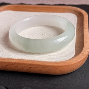 May include: A translucent jade bangle bracelet with a pale green hue. The smooth, polished surface reflects light, showcasing the natural variations in the stone. The bracelet is displayed on a wooden tray with a light-colored fabric lining.